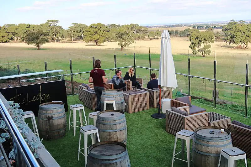 People seated at outdoor tables made from wooden barrels and wicker furniture at del Rios Winery, overlooking a wide landscape with trees and fields. The setting includes stools and a white umbrella, providing a scenic view of the countryside.